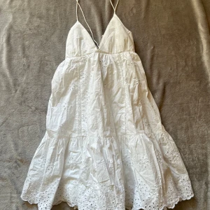 Vit sommarklänning från Zara XS - Superfin vit kortklänning från Zara med tunna axelband, v-ringning och broderade blommor. Klänningen har omlottknytning i ryggen och volangkant nertill. Perfekt för varma dagar och har en luftig passform i bomull. Har en reva på insidan men inget som syns när man har på sig den❤️