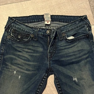 Mörkblå  jeans från True Religion - Säljer ett par mörkblå skinny jeans från True Religion med slitna detaljer och snygga kontrastsömmar. Jeansen har unika ficklock med knappar och klassisk logga bak. Perfekt för dig som gillar streetstyle och vill sticka ut.