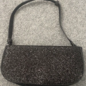 Glittrig svart axelväska från NA-KD - Säljer en snygg svart axelväska från NA-KD med glittrig yta och stilren form. Väskan har ett huvudfack med dragkedja och justerbar axelrem. Perfekt för att lyfta vilken outfit som helst med lite extra bling.
