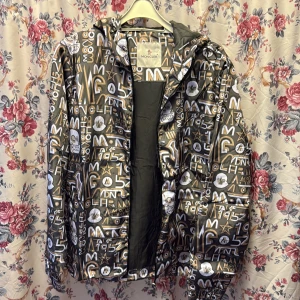 Moncler vindjacka med grafiskt mönster - Säljer en Moncler vindjacka med huva och dragkedja. Jackan har ett unikt grafiskt mönster i svart, vitt, guld och beige med Moncler-loggor och siffror. Tillverkad i glansig polyester och har en cool serietidningsdetalj på insidan. Perfekt för dig som vill sticka ut.