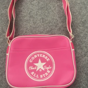 Rosa axelväska från Converse - Snygg rosa axelväska från Converse med vit logga och detaljer. Väskan har justerbar axelrem, dragkedja och ett stort huvudfack med innerficka. Perfekt för att bära med sig det viktigaste till skolan eller stan.har vissa synliga användningsområden men knappast syndliga 