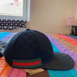 Svart Gucci keps med GG-mönster - Snygg svart keps från Gucci med klassiskt GG-monogram över hela kepsen. På sidan finns en grön och röd rand som ger den där ikoniska Gucci-looken. Justerbar rem baktill för perfekt passform. Perfekt accessoar för att lyfta din outfit.