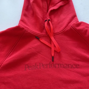 Röd hoodie från Peak Performance - Snygg röd hoodie från Peak Performance med matchande röda snören i huvan och diskret logga framtill. Klassisk passform och mjukt material som känns skönt mot huden. Aldrig använd.