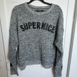 Grå stickad tröja från FB Sister - Säljer en gråmelerad stickad tröja från FB Sister med texten 'SUPERNICE' i svart framtill. Tröjan har lång ärm och coola svarta detaljer på ärmarna. Perfekt för dig som gillar en avslappnad och trendig stil.