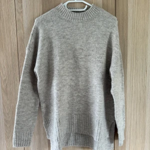 Beige stickad tröja från Vero Moda - Mysig beige stickad tröja från Vero Moda med ribbade muddar och högre krage. Tröjan har lång ärm och en lite längre, loose passform med slits på sidorna. Perfekt för lager-på-lager och chill höst- eller vinterstil.