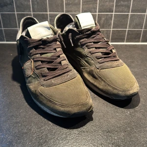 Philippe Model sneakers i olivgrönt - Snygga sneakers från Philippe Model i olivgrönt tyg och mocka med svarta snören och vita detaljer på hälen. Skorna har en klassisk sportig look med rund tå och platt sula. Perfekta för dig som gillar streetstyle och vill ha något unikt.