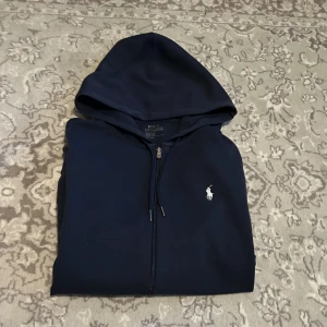 Mörkblå hoodie från Polo Ralph Lauren - Snygg hoodie från Polo Ralph Lauren med dragkedja och huva. Klassisk vit logga broderad på bröstet, långa ärmar och fickor framtill. Perfekt för en chill och stilren look. Tillverkad i mjuk bomullsmix för extra komfort.