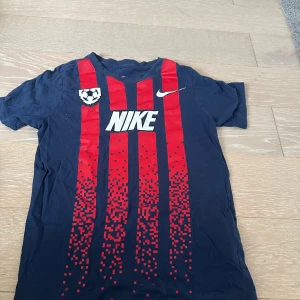 Blå och röd Nike fotbollströja, 137-147  - Snygg fotbollströja från Nike i mörkblått med röda vertikala ränder och pixelmönster framtill. Stor Nike-logga i vitt på bröstet och ett litet klubbmärke. Tillverkad i mjuk bomull, perfekt för träning eller match.