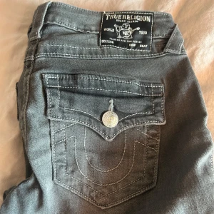 True Religion Joey grå flare jeans - Snygga grå jeans från True Religion, modell Joey Low Rise Flare. Byxorna har låg midja, utsvängda ben och klassiska detaljer som stora bakfickor med lock och silverfärgade knappar. Tydliga kontrastsömmar och mjukt denimtyg. Säljes då dem ej används, använda ca 2 gånger så inga defekter💓 storlek 27! Köpta på Nelly i Stockholm för ca 1200kr!