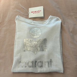 Vit t-shirt från Marant Etoile - Vit t-shirt från Isabella  Marant Etoile med ikoniskt silvertryck framtill. Klassisk rund halsringning och korta ärmar. Tillverkad i mjuk bomull, och aldrig använd som i nyskick 