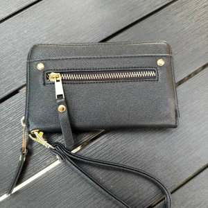 Svart plånbok med gulddetaljer - Snygg svart plånbok med struktur och guldfärgade detaljer. Har dragkedja runtom, ett yttre fack med dragkedja och flera kortfack inuti. Praktisk handledsrem gör den enkel att bära med sig. Perfekt för att hålla koll på kort, pengar och smågrejer. Aldrig använd😊