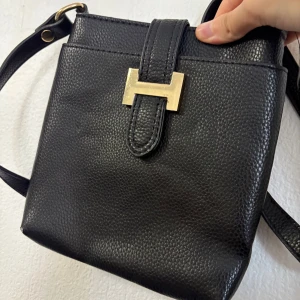 Svart axelväska i skinn med gulddetalj - Snygg svart axelväska i präglat skinn med justerbart axelband och en stilren guldfärgad H-detalj framtill. Väskan har en klassisk fyrkantig form och stängs med ett lock. Perfekt för dig som gillar minimalistisk och tidlös design.