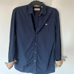 Mörkblå skjorta från Burberry - Snygg mörkblå skjorta från Burberry med klassisk krage och knappar framtill. Ikoniskt Burberry-mönster syns på insidan av ärmsluten. Liten broderad logga på bröstet. Perfekt för dig som gillar stilrena och exklusiva plagg. Äkta 