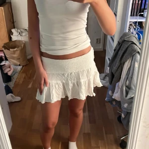 Vit volangkjol med smock - Supersöt vit kortkjol med bred smock i midjan och volangig nederdel. Kjolen är lätt och luftig, perfekt för varma dagar. Den har en feminin och flowy känsla som passar till många olika stilar.