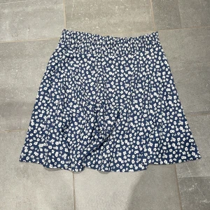 Blommig blå kortkjol med resårmidja - Supersöt blå kortkjol med vita blommor och bekväm resår i midjan. Kjolen har en lätt utställd form och är perfekt för varma dagar. Materialet känns mjukt och luftigt, vilket gör den skön att bära hela dagen. Går att diskutera pris