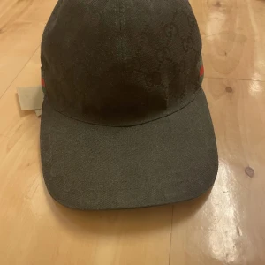 Svart keps från Gucci med GG-mönster - Svart keps från Gucci med klassiskt GG-monogram över hela kepsen. Sidorna har gröna och röda ränder som är typiska för märket. Justerbar rem baktill i skinn. Snygg och stilren keps som lyfter vilken outfit som helst.