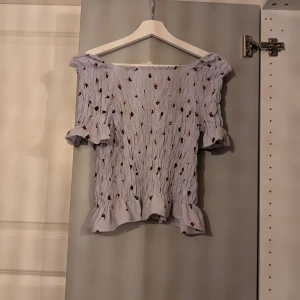 Ljuslila blommig offshoulder blus - Supersöt ljuslila blus med små blommor, smockad över hela kroppen och volangdetaljer vid ärmslut och nederkant. Offshoulder-modell med korta ärmar, perfekt för en somrig look. Toppens material känns lätt och luftig.