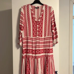 Rödvit mönstrad kortklänning - Superfin kortklänning från Montego i vitt och rött med grafiskt mönster. Klänningen har v-ringning, trekvartsärm och en luftig passform. Perfekt för dig som gillar boho-vibes och vill sticka ut med coola detaljer.