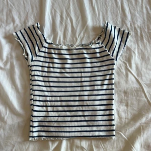 Randig ribbad topp med volangkant - Superfin vit topp med marinblå ränder och ribbad struktur. Modellen är croppad med korta ärmar och har söt volangkant vid halsringning och nederkant. Perfekt för en chill sommarlook.