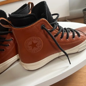 Bruna Converse All Star skinnsneakers - Snygga höga Converse All Star sneakers i brunt skinn med svarta snören och svart plös. Klassisk vit sula och tydlig Converse-logga på sidan. Perfekta för dig som vill ha en stilren och tidlös look med en twist. Passar till både jeans och streetwear.
