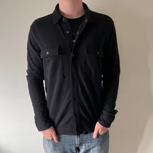 Svart overshirt med knappar från selected home - Stilren svart overshirt med klassisk krage och två stora bröstfickor med lock och knappar. Långärmad modell i mjukt material, perfekt att bära öppen eller stängd. Enkel och clean look som passar till jeans eller andra byxor.