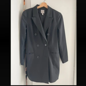 Svart kavajjacka med dubbelknäppning - Snygg lång svart kavajjacka med klassisk dubbelknäppning och långa ärmar. Kavajen har två fickor framtill och en rak, något längre passform.