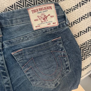 True Religion bootcut jeans blå, stl 26 - Säljer ett par lågmidjade blå jeans från True Religion i bootcut. Ljusrosa sömmar och broderad bakficka. 