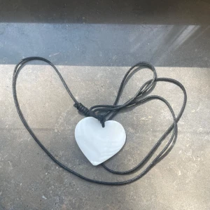 Halsband med vitt hjärthänge - Snyggt halsband med ett vitt hjärta i blankt material, upphängt på en svart justerbar läderrem. Hjärtat har en mjuk, rundad form och ger en stilren och enkel look.