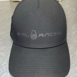 Sail Racing Keps - Svart keps från Sail Racing med en stilren och diskret logga framtill. Kepsen har en böjd skärm och ett justerbart metallspänne baktill för perfekt passform. Tillverkad i ett lätt och syntetiskt material som ger en modern och sportig look. Mycket fint skick. Skicka gärna ett DM vid frågor! Pris kan diskuteras vid snabb affär. 