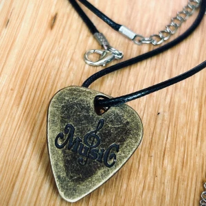 Vintage-halsband med plektrum i brons! - Vintage-halsband med plektrum i brons med en G-klav & ”Music” ingraverat. Halsband med tunn läderrem medföljer. Mått plektrum: 2,5 x 3 cm. Kedja: 45 cm (+6). I nyskick!