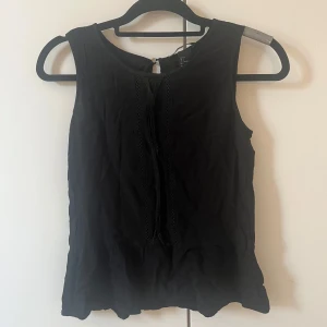 Svart ärmlös blus med spetsdetaljer - Svart ärmlös blus från H&M med fina spetsdetaljer längs framsidan och en liten knapp i nacken. Toppen har en lätt volangkant nedtill och är tillverkad i mjuk bomull.