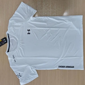 Vit Under Armour t-shirt HeatGear - Vit t-shirt från Under Armour med svarta loggor på bröstet, ärmen och nedtill. Tillverkad i lätt och stretchig polyester, perfekt för träning. Modellen är kortärmad med rund hals och har en clean, sportig look.helt oanvänd möts upp i goteborg
