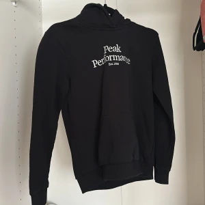 Svart hoodie från Peak Performance - Svart hoodie från Peak Performance med vit logga på bröstet.  Mjuk bomullskänsla och ribbade muddar vid ärmslut och nederkant. 