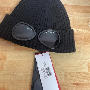 Svart  C.P. Company mössa - Svart ribbstickad mössa från C.P. Company med ikoniska inbyggda goggles framtill. Mössan har bred uppvikt kant och är tillverkad i mjuk merinoull. Perfekt accessoar för att sticka ut och hålla värmen under kalla dagar.