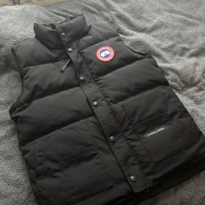 Svart dunväst från Canada Goose - Säljer en svart dunväst från Canada Goose med klassisk logga på bröstet. Västen har hög krage, knappar och dragkedja framtill samt två stora fickor. Perfekt för kyliga dagar och riktigt snygg till många outfits.