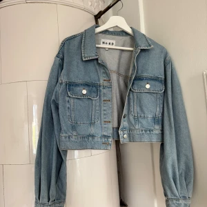 Croppad jeansjacka från NA-KD - Säljer en croppad jeansjacka från NA-KD i ljusblå denim. Jackan har långa puffärmar och två bröstfickor med knappar 