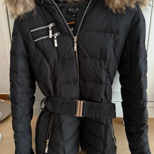 Jacka från Hollies - Säljer nu min snygga svarta dunjacka från Hollies med fuskpäls på huvan, markerad midja med brett bälte och flera dragkedjedetaljer. Inte använt den på flera år, så tänkte att den kan få en ny ägare