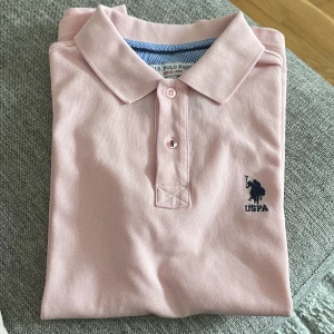 Ljusrosa pikétröja från U.S. Polo Assn. - Säljer en ljusrosa pikétröja från U.S. Polo Assn. i slim fit. Tröjan har korta ärmar, klassisk krage och knäppning framtill. Broderad logga i mörkblått på bröstet. Perfekt för en stilren och sportig look.