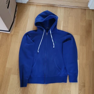 Blå hoodie från Polo Ralph Lauren - Säljer en klassisk blå hoodie från Polo Ralph Lauren med dragkedja och vit snörning i huvan. Tröjan har två fickor framtill och är tillverkad i mjukt material. Perfekt för en avslappnad stil.