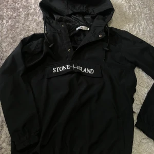 Svart vindjacka från Stone Island - Svart vindjacka med huva från Stone Island. Jackan har en broderad logotyp framtill och dragsko vid huvan. Perfekt för blåsiga dagar och enkel att matcha med olika outfits.