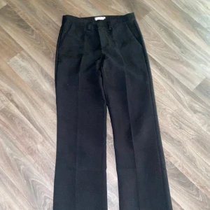 Svarta kostymbyxor med raka ben - Säljer ett par klassiska kostymbyxor från Nelly med klassisk rak passform med en liten utsvängning och pressveck. Byxorna har hällor för bälte och stängs med dragkedja och hyska. Perfekta för en stilren look. Burit de en gång och är i nytt skick. 🖤