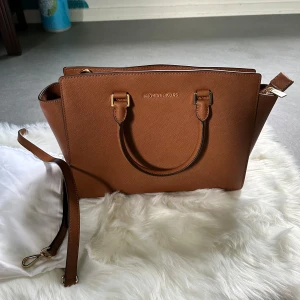 Brun handväska från Michael Kors - Säljer en snygg brun handväska från Michael Kors, modell Selma. Väskan är tillverkad i läder och har både handtag och avtagbar axelrem. Perfekt storlek för att få plats med allt du behöver. Guldiga metalldetaljer och stilren design.