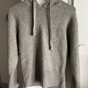 Grå stickad hoodie - Mysig grå stickad hoodie med långa ärmar och ribbade muddar. Tröjan har en huva med dragsko och är perfekt för kyliga dagar. Enkel och stilren design som passar till det mesta.