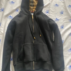Svart hoodie med dragkedja från Burberry - Svart hoodie från Burberry med klassiskt rutigt foder i huvan. Jackan har dragkedja i guld, två stora fickor framtill och justerbar huva med snören. Perfekt för en stilren och avslappnad look.
