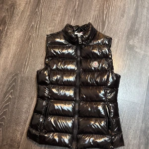 Svart dunväst från Moncler - Snygg svart dunväst från Moncler med glansig finish och hög krage. Västen har dragkedja framtill och två fickor med dragkedja. Klassisk Moncler-logga på bröstet och serietidningsetikett på insidan.