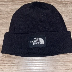 Svart mössa från The North Face - Svart ribbstickad mössa från The North Face med klassisk logotyp framtill. Tillverkad i mjuk polyesterblandning med lite stretch för skön passform. Perfekt för kyliga dagar. 9/10 skick. Kvitto ingår inte