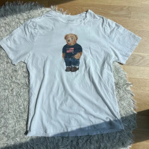 Vit t-shirt med björntryck från Polo Ralph Lauren - Vit t-shirt från Polo Ralph Lauren med ett tryck av en björn på framsidan som bär en tröja med amerikansk flagga. Trycket e lite slitet därav priset, men ja tycker nästan bara det blir snyggare. Står ingen storlek i den men den sitter som en Medium. Höra gärna av dig om du har några frågor!