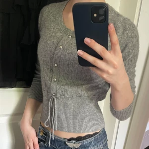 Grå stickad tröja i 100% kashmir - Superfin grå stickad tröja i 100% kashmir. Koftan har knappar framtill, ribbade muddar och ett snöre i midjan för en bättre passform. Croppad modell med trekvarts ärm. Använd en gång.