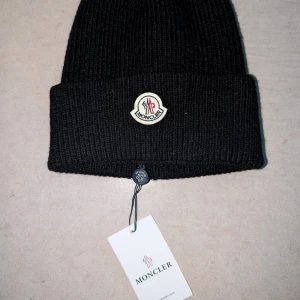 Svart ribbstickad mössa från Moncler - Svart ribbstickad mössa från Moncler med klassisk Moncler-logga framtill. Mössan har uppvikt kant och är tillverkad i ett mjukt och värmande material, perfekt för kyliga dagar.