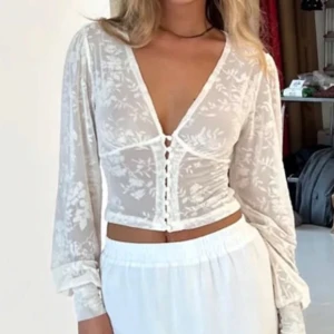 Vit blus med knappar från Venderbys - Säljer en romantisk vit blus från Venderbys med v-ringning, genomskinligt blommigt tyg och dekorativa knappar framtill. Blusen har långa ärmar och en figurnära passform, perfekt för att lyfta vilken outfit som helst.